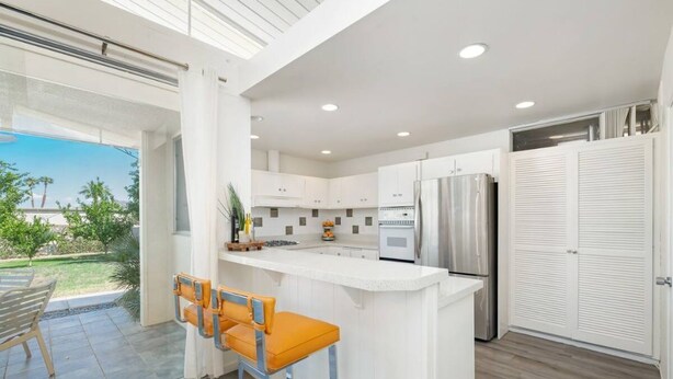 Una cocina moderna con armarios blancos, una isla grande con taburetes de bar, un refrigerador y vistas a un patio con mobiliario exterior.