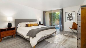 Un dormitorio moderno con una cama grande, una mesita de noche de madera, un sillón de cuero y vistas al exterior con vegetación.