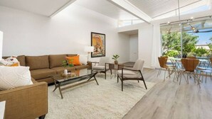 Una sala de estar moderna con sofá, mesa de centro y sillas, una mesa con cubierta de vidrio y vistas a áreas verdes.