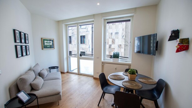 A modern living room with a sofa, a dining table with chairs, a TV mounted on the wall, and large windows with a view of buildings. - Luxury Apartment With Balcony (Como)