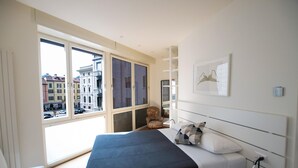 A bedroom with a bed, a chair, a window with blinds, and a city view. - Luxury Apartment With Balcony (Como)
