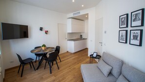 A modern living room with a grey sofa, a dining table with chairs, a flat-screen TV mounted on the wall, and a kitchen area with white cabinets and a countertop. - Luxury Apartment With Balcony (Como)