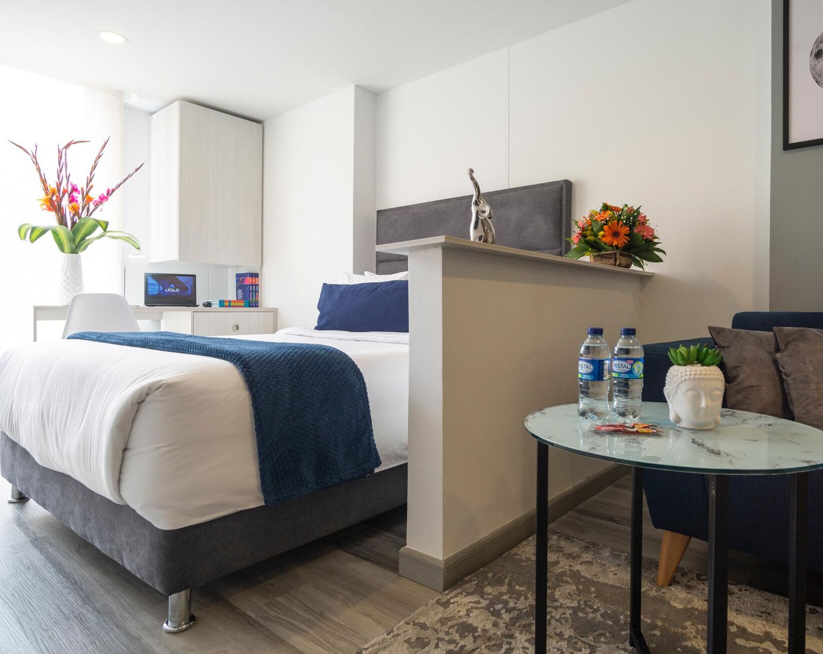 Una habitación de hotel moderna con cama, mesita de noche y una pequeña mesa redonda con botellas de agua y una planta.
