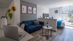Un salón moderno con un sofá azul, una mesa de centro de cristal y un ordenador portátil sobre un escritorio. Hay cuadros enmarcados en la pared y un ventanal con vistas a la ciudad.