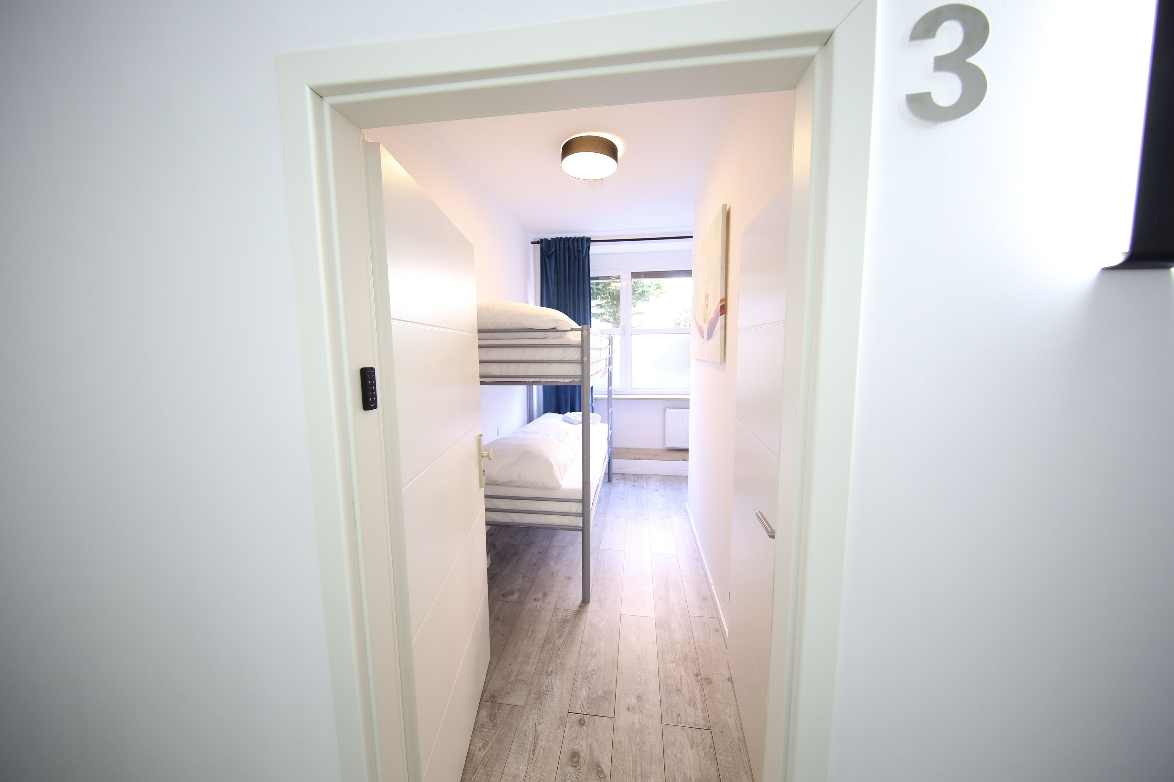 Un couloir étroit, avec un lit superposé au fond, le chiffre « 3 » peint sur le mur et une fenêtre avec des rideaux.