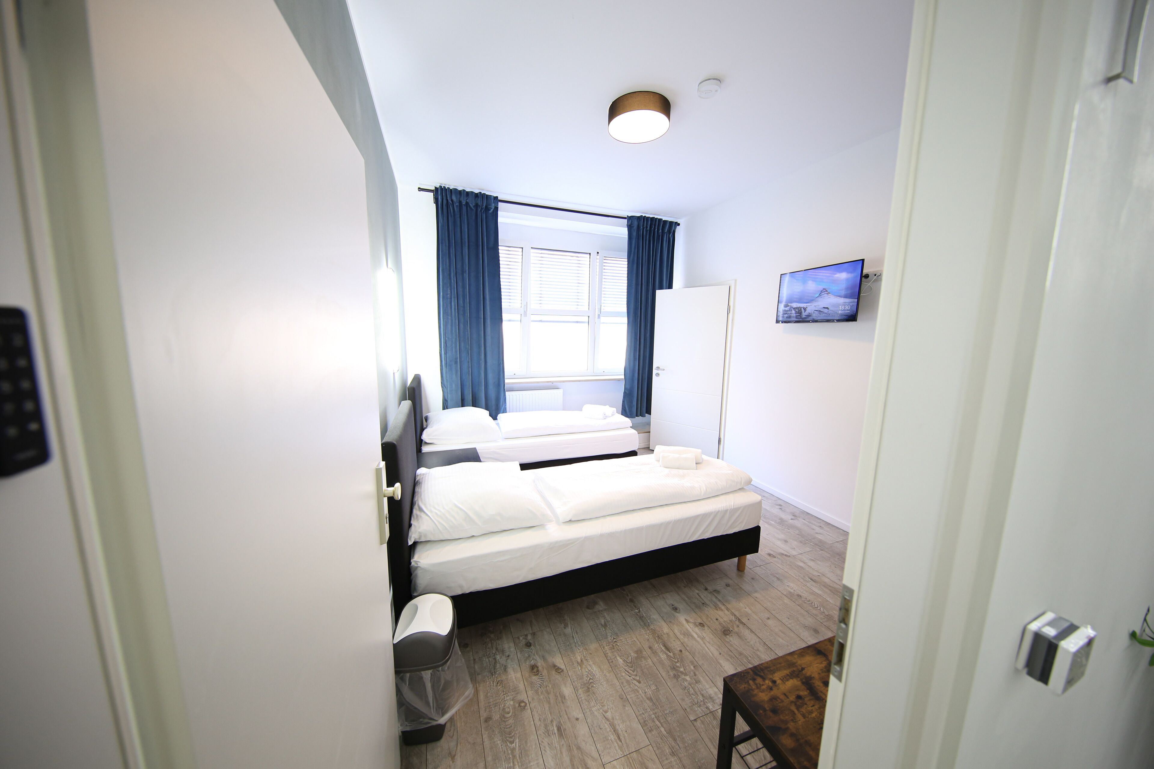A bedroom with two beds, a window with curtains, a TV on the wall, and a wooden floor.