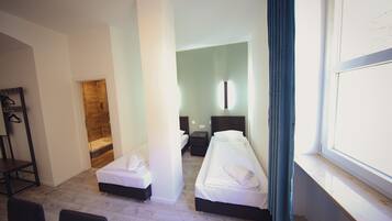 A hotel room with two single beds, a wooden floor, a large window with curtains, a desk, and a wardrobe.