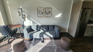 Ein modernes Wohnzimmer mit einem grauen Sofa, einem Glastisch und zwei Hocker. Es gibt einen Schreibtisch mit einem Stuhl und einer Lampe. Zwei gerahmte Kunstwerke sind an der Wand angebracht.