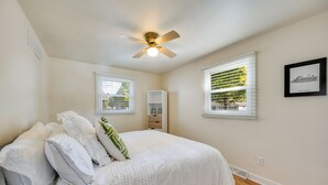 A bedroom with a bed, white bedding, a ceiling fan, two windows with blinds, a white wardrobe, and a framed picture on the wall.