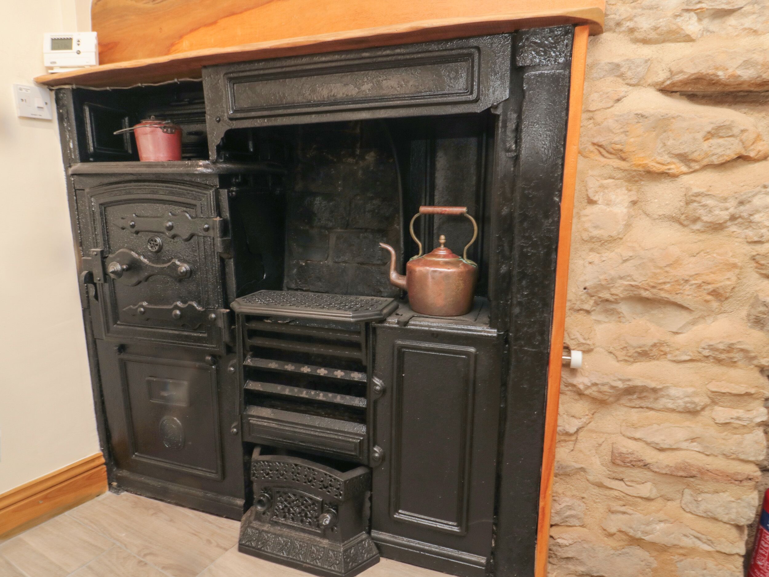 A black, ornate fireplace with a wooden mantel, a red pot on the left, and a copper kettle on the right.