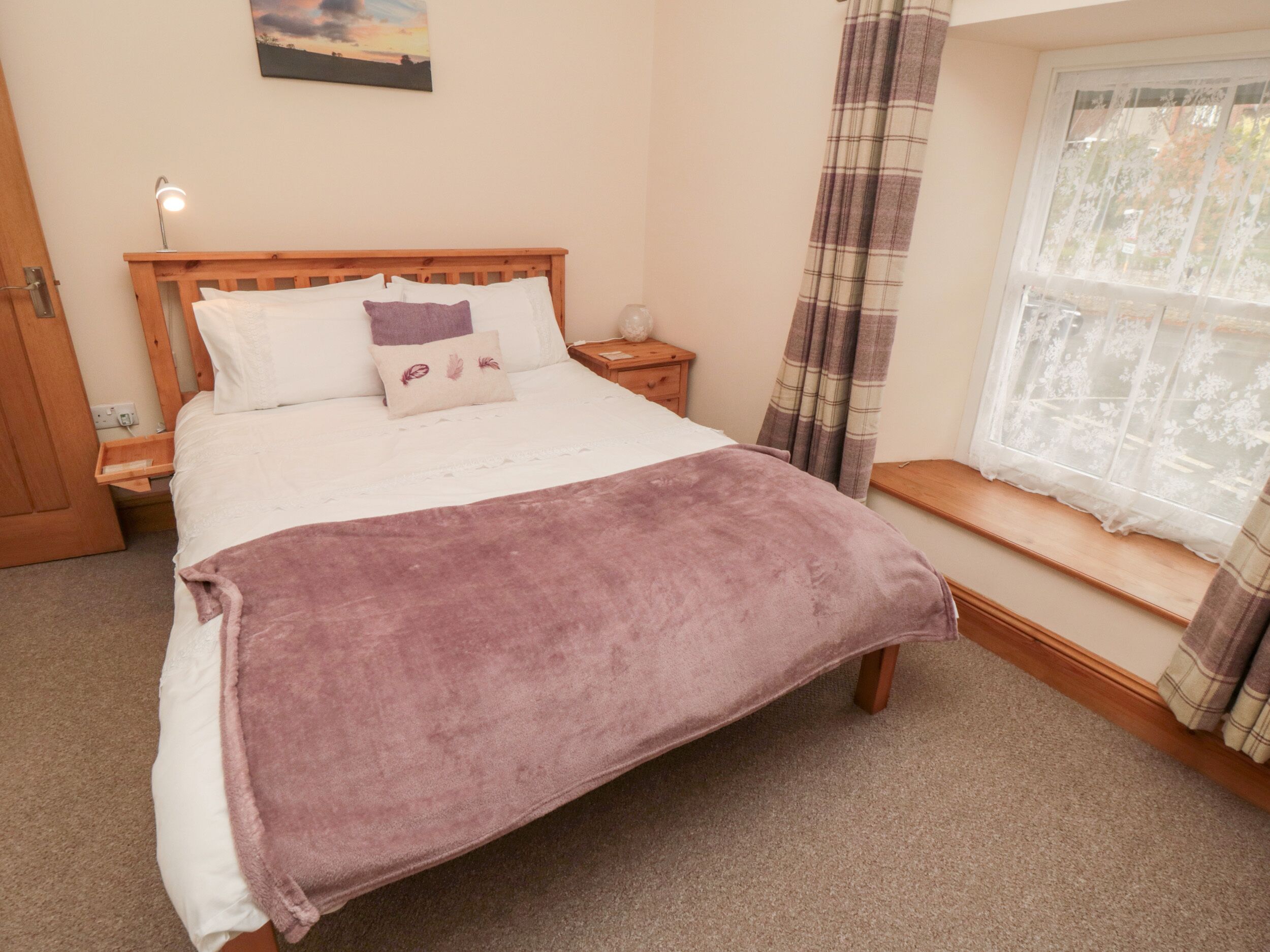A neatly made bed with a purple blanket, a bedside table, a nightstand, a window with curtains, and a painting on the wall.