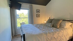 Un dormitorio con una cama, una ventana con cortinas, una lámpara en el techo y dos cuadros en la pared.