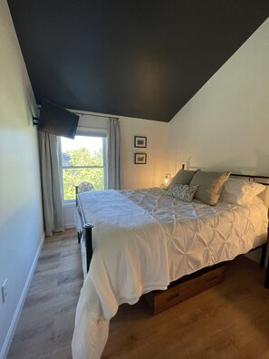 Une chambre à coucher avec un lit, une fenêtre avec des rideaux, un luminaire au plafond et deux tableaux encadrés accrochés au mur.