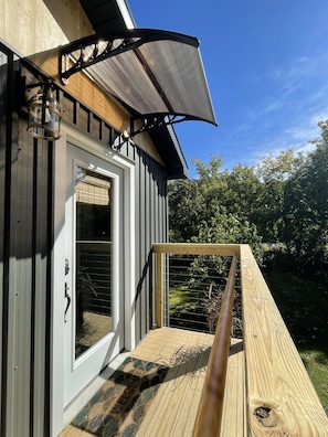 Une terrasse en bois avec une rambarde en métal, une porte dotée d’une fenêtre et un auvent noir.