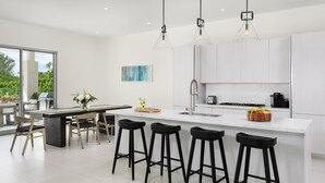 Une cuisine moderne avec des meubles blancs, un îlot central doté de tabourets de bar, un éclairage suspendu et une vue sur l’extérieur.