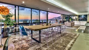 Un salón moderno con una gran mesa de madera, sillas transparentes, un sofá y vistas a la ciudad y montañas a través de amplios ventanales.