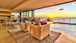 Una sala de estar moderna con un gran ventanal con vista a una piscina y al océano al atardecer.