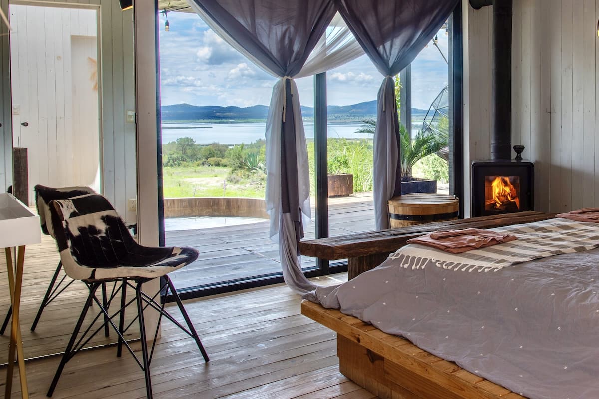 Quarto com uma cama grande, um banco de madeira, uma cadeira e vista para um lago e montanhas através de grandes janelas.