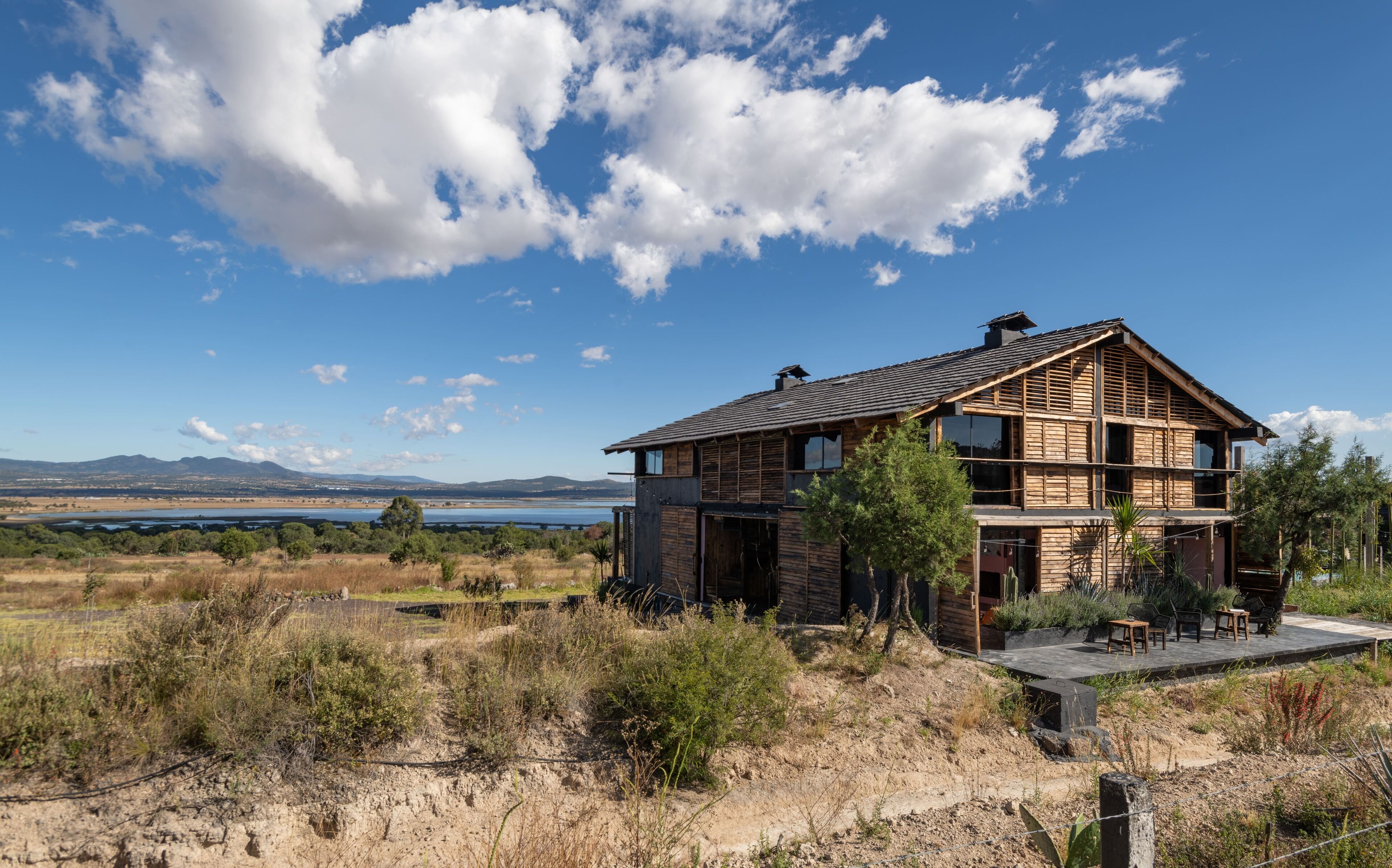 A two-story wooden house with a balcony, surrounded by dry grass and shrubs, with a lake and mountains in the background.
