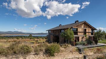 A two-story wooden house with a balcony, surrounded by dry grass and shrubs, with a lake and mountains in the background.