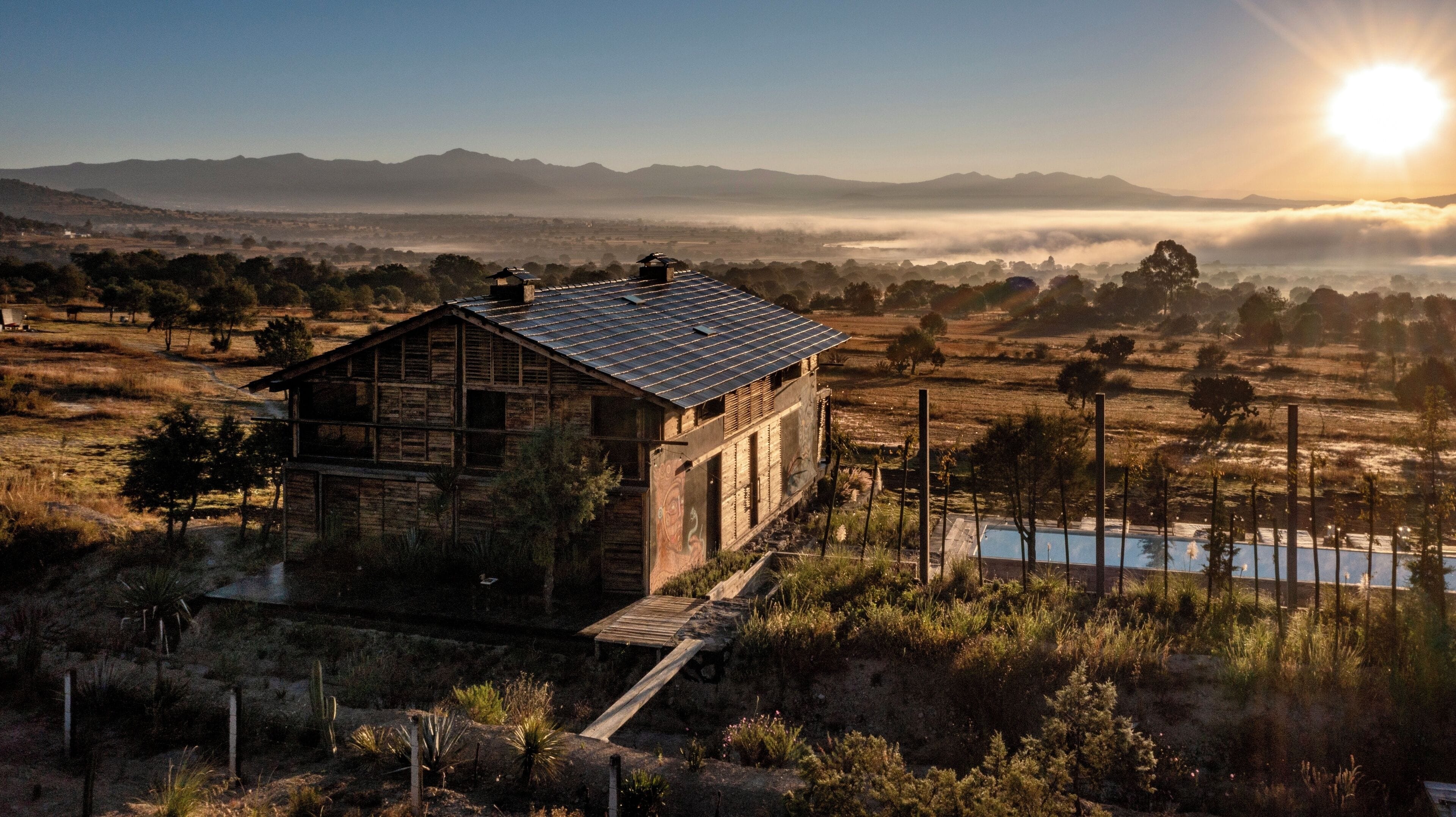 A rustic cabin with a metal roof, surrounded by a dry landscape and a body of water.