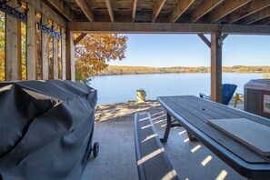 An outdoor seating area with a covered picnic table and a grill, overlooking a calm lake with autumn foliage.
