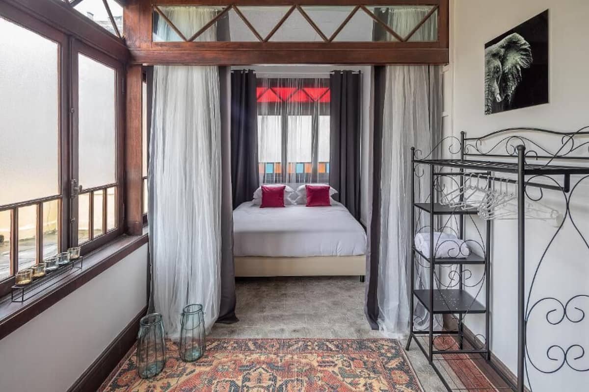 A bedroom with a bed, curtains, a window, a metal rack, and a patterned rug.