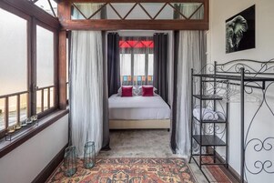 Un dormitorio con una cama, cortinas, un ventanal con vistas, un estante metálico y una alfombra estampada.