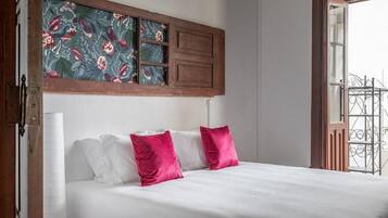Una cama con almohadas blancas y rosas, un cabecero de madera con un panel floral y vistas a un balcón con barandilla metálica.