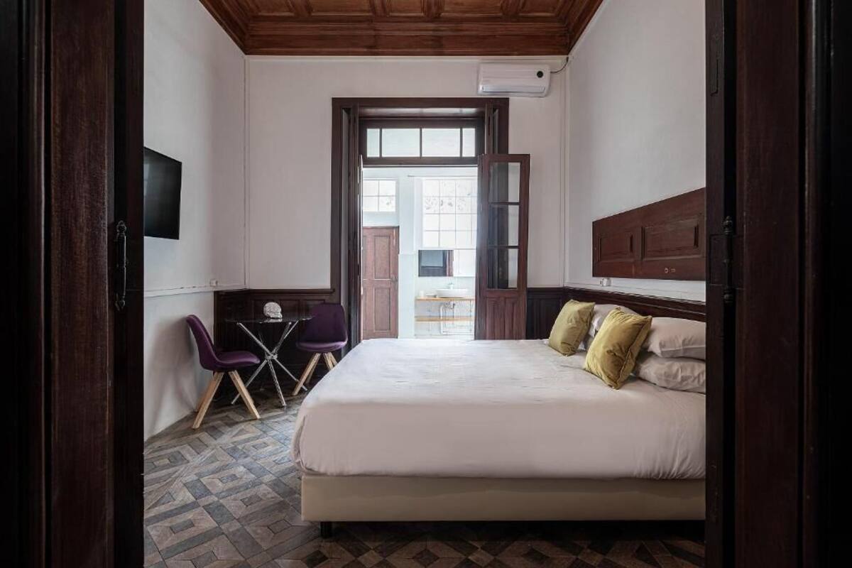 A bedroom with a bed, a desk, and a chair. There is a window and a door leading to another room.