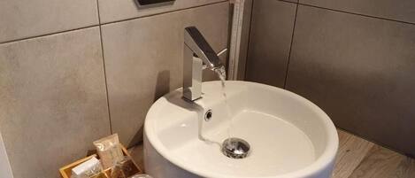 Un baño con un lavamanos redondo blanco, un grifo y un espejo encima. Hay una bandeja de madera con artículos de aseo en el borde del lavamanos.