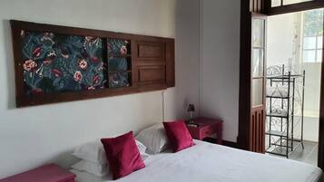 A bedroom with a bed, two nightstands, a red chair, a decorative wall panel, and a view of the outside.