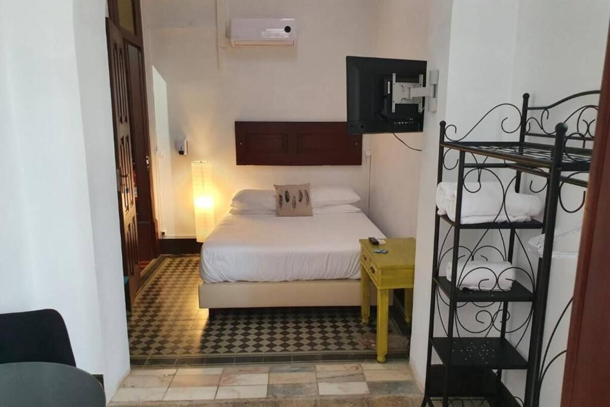 A small, compact room with a bed, a bedside table, a wall-mounted TV, and a metal shelf unit.