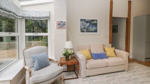 Un salon comprenant un canapé, un fauteuil et une petite table avec une lampe. Il y a une fenêtre donnant sur un jardin et un tableau accroché au mur.