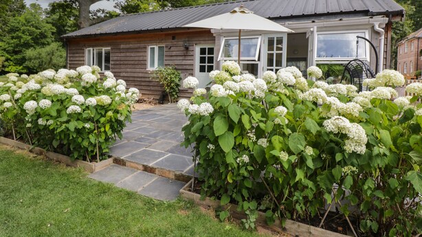 Une maison en bois dotée d’un patio carrelé, entourée d’une végétation luxuriante et de buissons d’hortensias blancs.