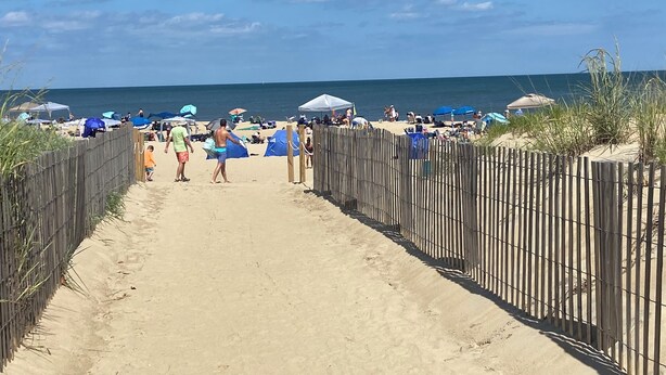 Ein Sandweg am Strand, der zum Meer führt, mit Liegestühlen und Menschen.