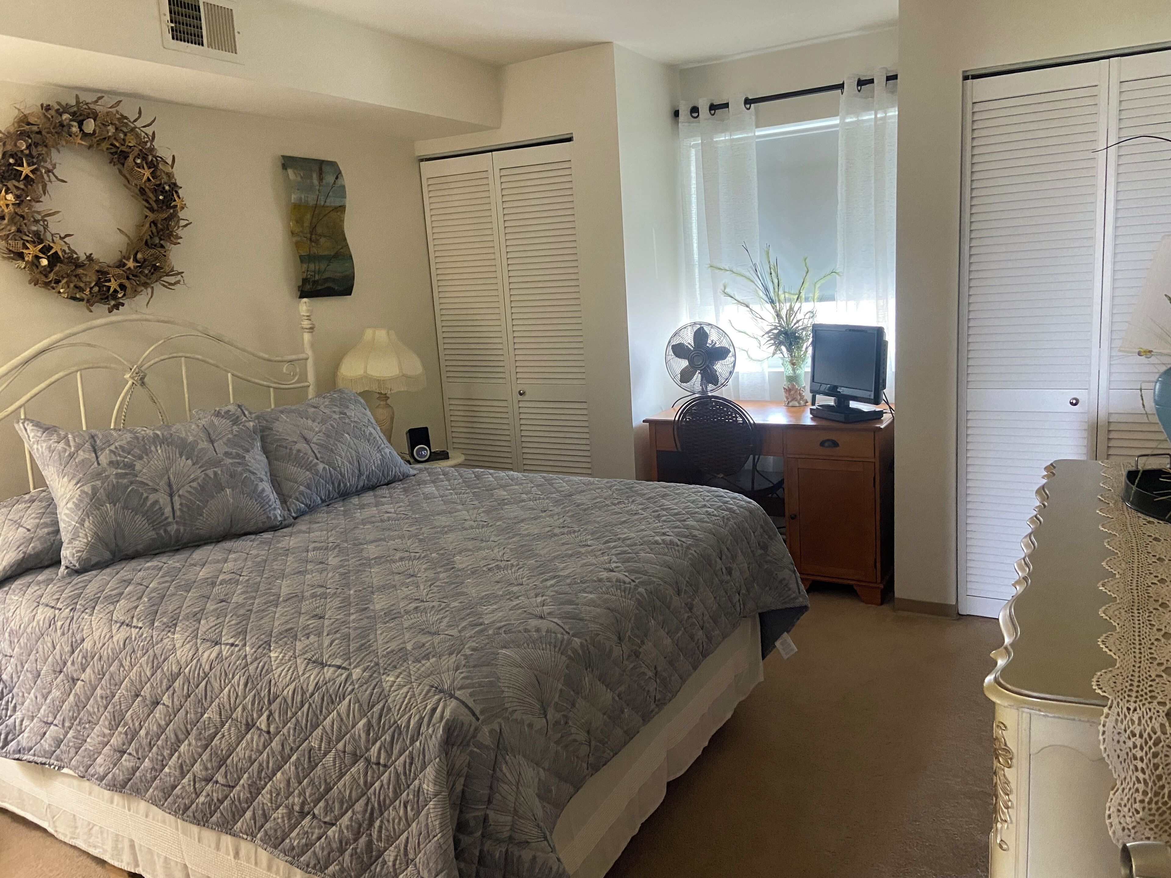 A bedroom with a bed, a desk with a computer, a fan, and a window with curtains.