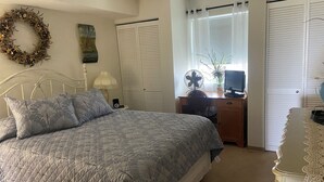 Ein Schlafzimmer mit einem Bett, einem Schreibtisch mit Computer, einem Ventilator und einem Fenster mit Vorhängen.