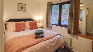 A bedroom with a bed, a nightstand, a lamp, a window with curtains, a radiator, and a mirror. - Woodland Retreat (Windermere)
