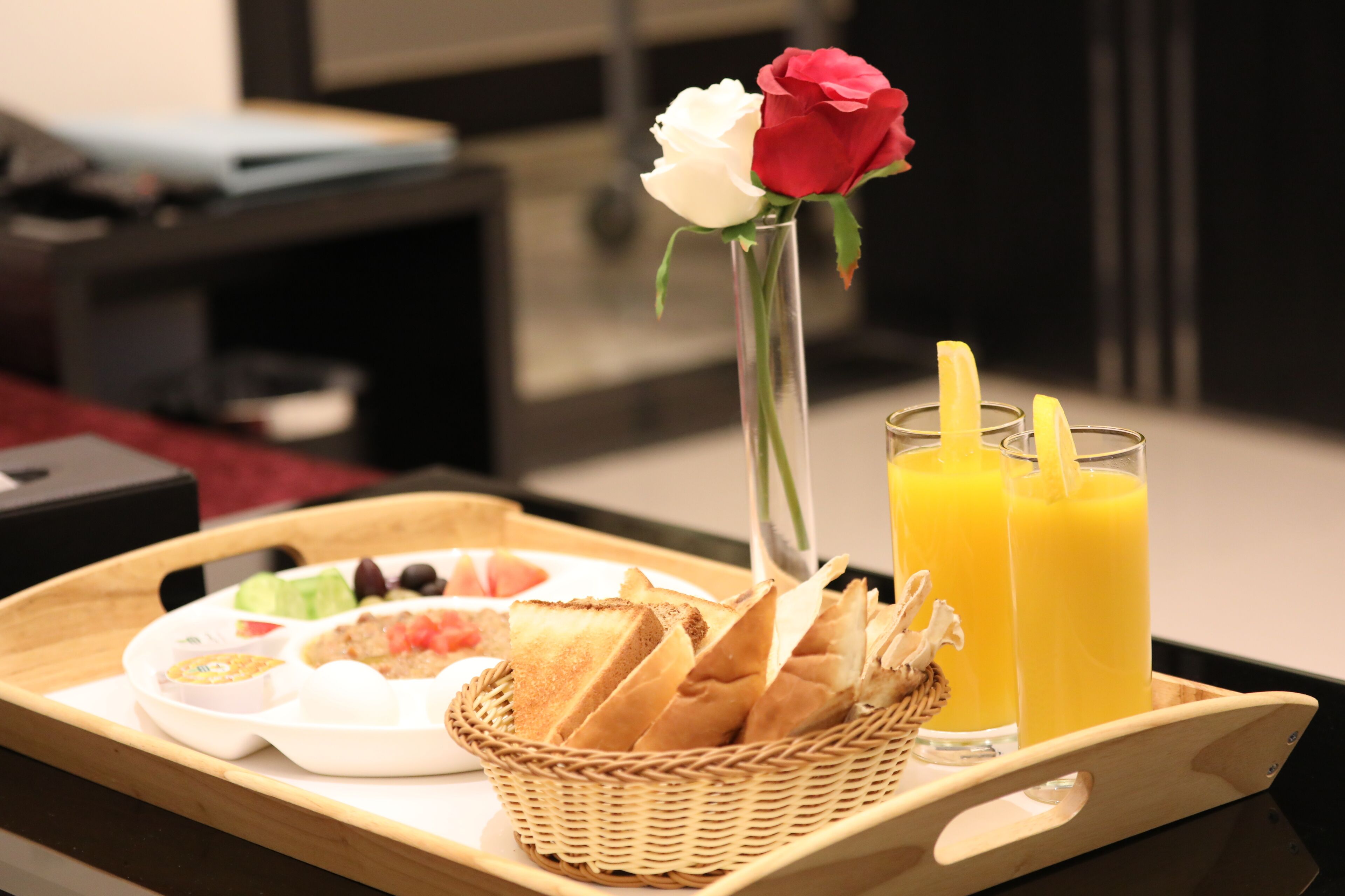 Ein Frühstücksbrett mit Toast, Obst und Saft, eine Vase mit einer Rose und ein Korbwesen.