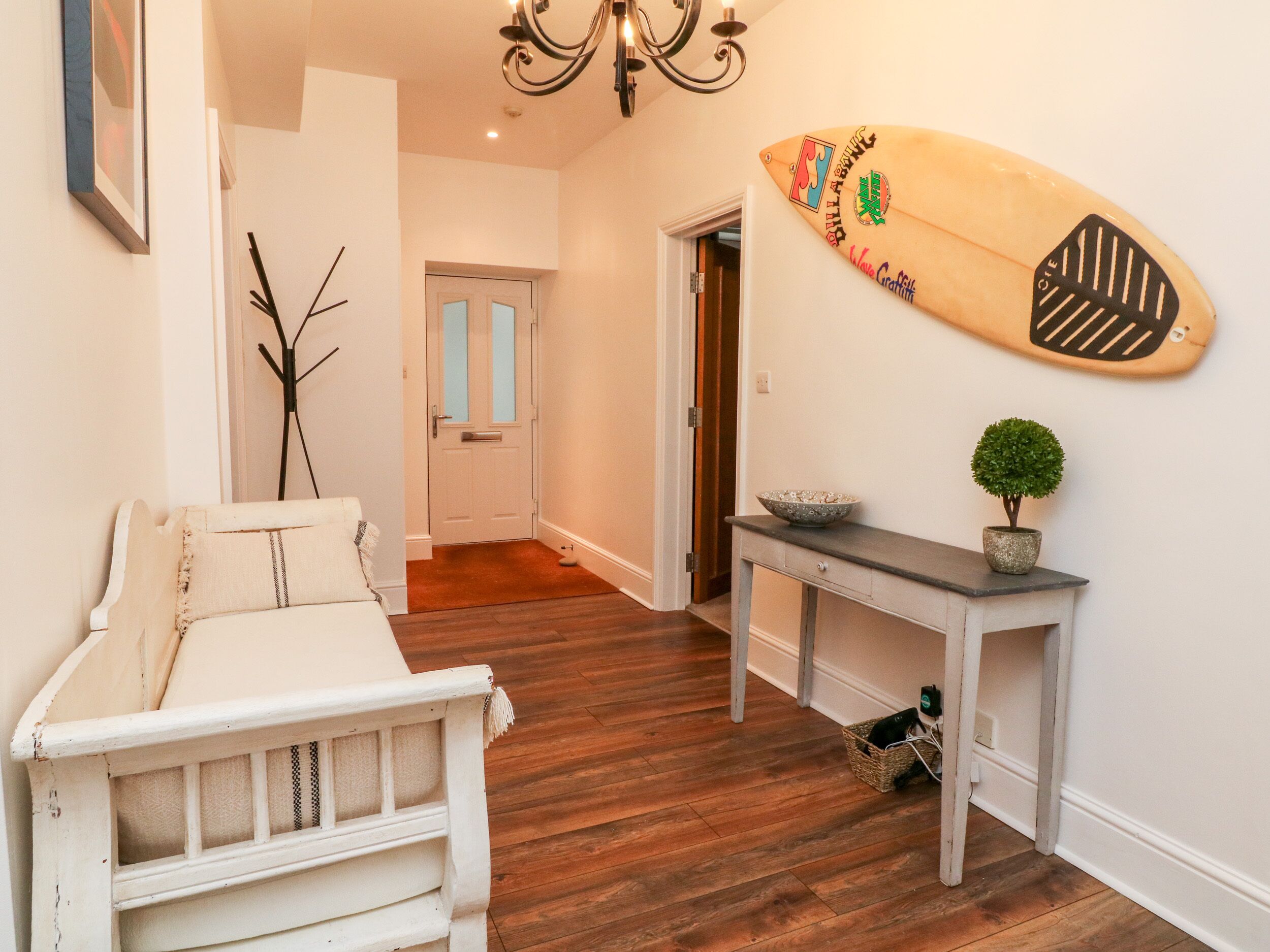 A hallway with a white bench, a wooden floor, a wall-mounted surfboard, a small potted plant, and a door with frosted glass panels.