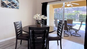 Comedor con una mesa redonda, cuatro sillas, un florero con flores blancas, un ventilador de techo y vistas a una piscina a través de un ventanal.