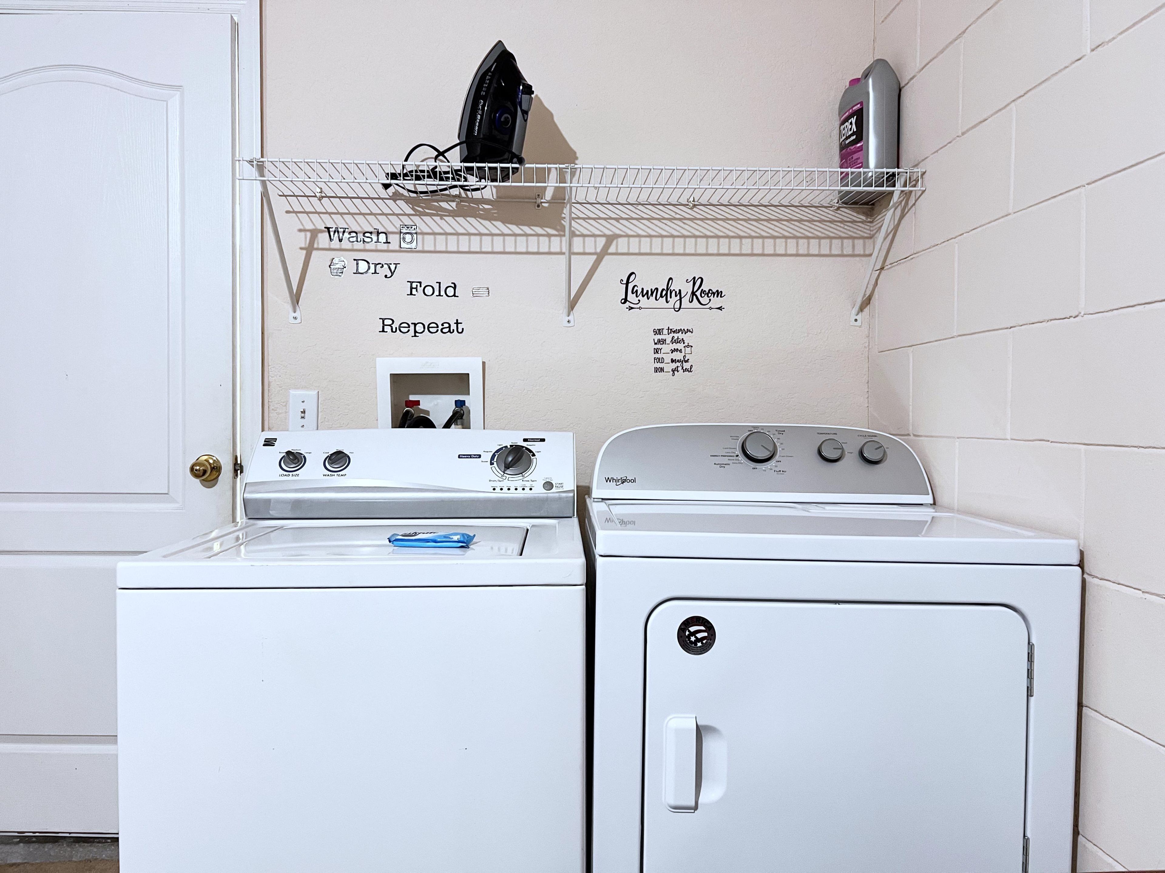 A laundry room with a front-loading washer and dryer, a drying rack, and a wall-mounted shelf with laundry supplies.