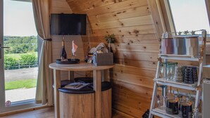 A cozy cabin interior with a wooden ceiling, a television mounted on the wall, a small table with a bottle and glass, and a shelf with jars and mugs. - Joshua's Den - UK40975 (Near Swansea)