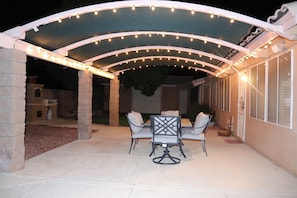 Un patio exterior con zona de estar techada, luces colgantes y una pared de piedra.