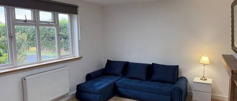 A modern living room with a sectional sofa, a radiator, a window with blinds, a lamp, and a mirror on the wall.