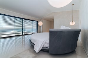 Un dormitorio moderno con una cama curvada, ventanas amplias con persianas horizontales y dos lámparas colgantes.