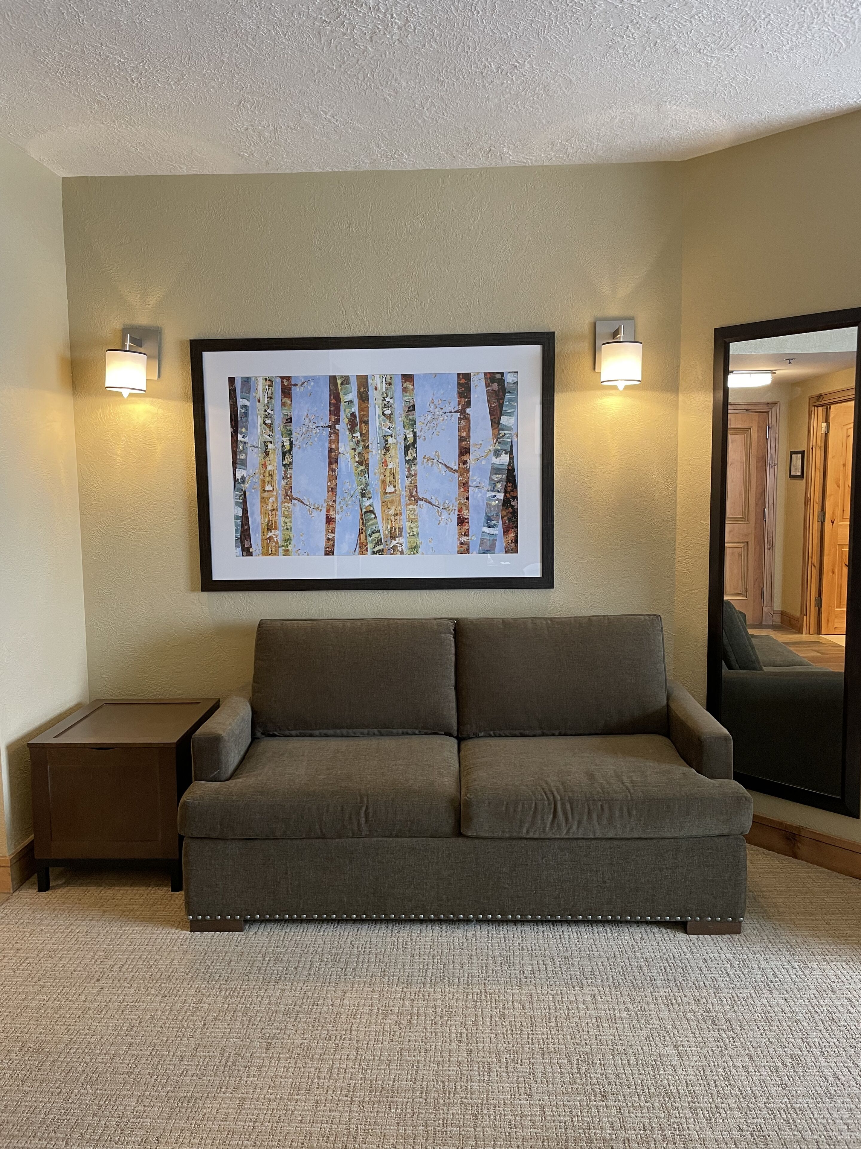 A neutral-toned living room with a gray sofa, a framed abstract painting, and two wall-mounted light fixtures.