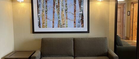 A neutral-toned living room with a gray sofa, a framed abstract painting, and two wall-mounted light fixtures.
