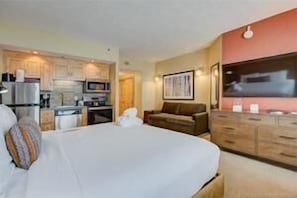 A hotel room with a bed, a sofa, a TV, a kitchen area, and a dining table.